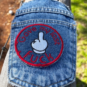 May include: A blue denim pocket featuring a circular embroidered patch. The patch has a red border and contains a white cartoon hand gesture of a middle finger. The red embroidered text reads "I gave my last FUCK" around the central graphic.