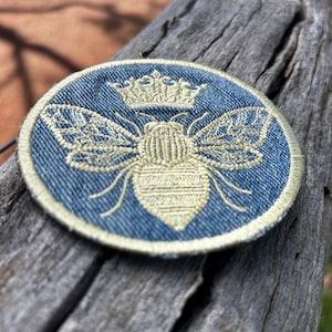 May include: A circular denim patch featuring an embroidered light yellow bee design topped with a small crown. The patch has a stitched border in a matching light yellow thread, creating a distinct contrast against the blue denim fabric background.
