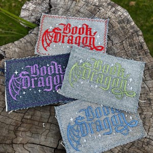 May include: Four rectangular denim patches with frayed edges, each featuring an embroidered dragon silhouette and the text "Book Dragon" in a gothic font, accented by small white stars. The embroidery colours are red, purple, lime green, and blue.