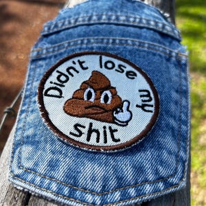 May include: A circular embroidered patch attached to a blue denim pocket. The patch features a brown cartoon poop character with a grumpy expression, giving the middle finger. The text "Didn't lose my shit" is stitched in black around the edge.