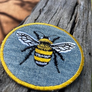 May include: A circular embroidered patch featuring a bumblebee with yellow and black stripes, white wings with black outlines, and black legs. The background of the patch is a light blue fabric, and the entire edge is finished with a yellow border.