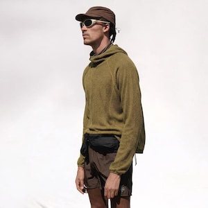 Puede incluir: Una persona lleva una gorra marrón, gafas de sol y un suéter con capucha verde. También lleva pantalones cortos marrones y una riñonera negra. El fondo es blanco.