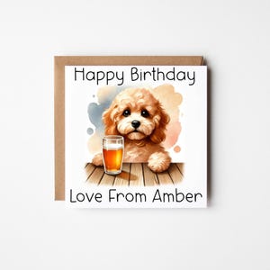 May include: A square birthday card featuring a light brown dog illustration holding a pint of beer. The card has the text "Happy Birthday" and "Love From Amber". The background has watercolor splashes and the card is set on a white background.