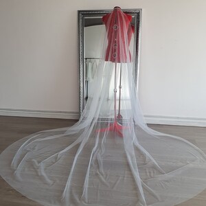 May include: A white tulle wedding veil with a long train, displayed on a mannequin. The veil is made of soft, delicate fabric and has a classic design.