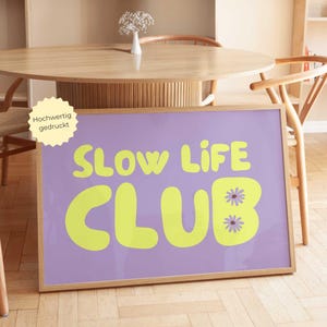 May include: A framed poster with the words "SLOW LIFE CLUB" in bright yellow, set against a lavender background. The poster includes two daisy graphics and is in a light wood frame. The image is displayed in a room with wooden furniture.