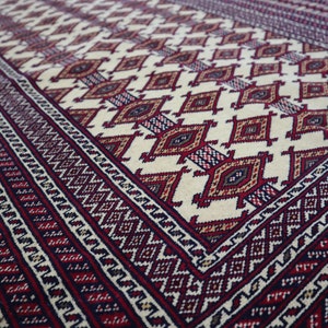 May include: A close-up of a traditional rug with a cream background and intricate red, blue, and brown geometric patterns. The rug has a border of repeating diamond shapes.