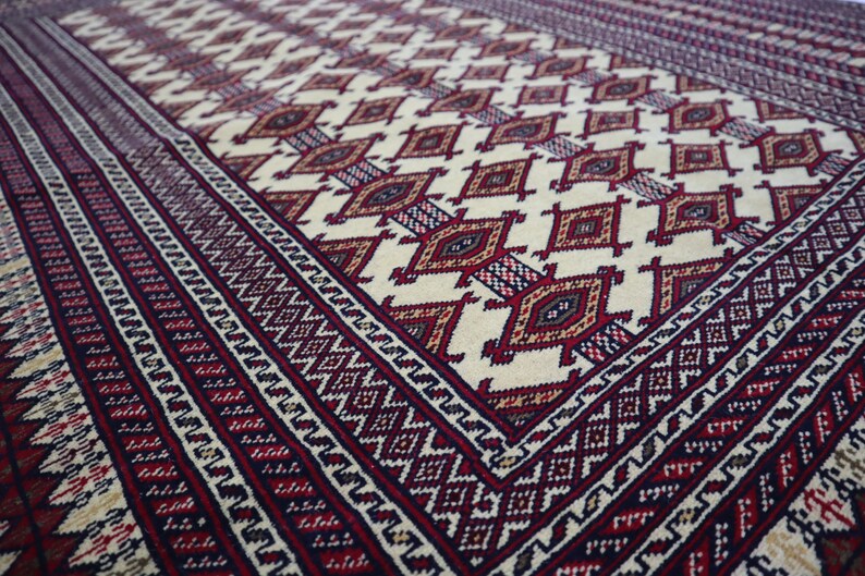 May include: A close-up of a traditional rug with a cream background and intricate red, blue, and brown geometric patterns. The rug has a border of repeating diamond shapes.