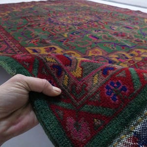 May include: A close-up of a green, red and yellow patterned rug with a fringed border. The rug is folded in half and being held by a hand.