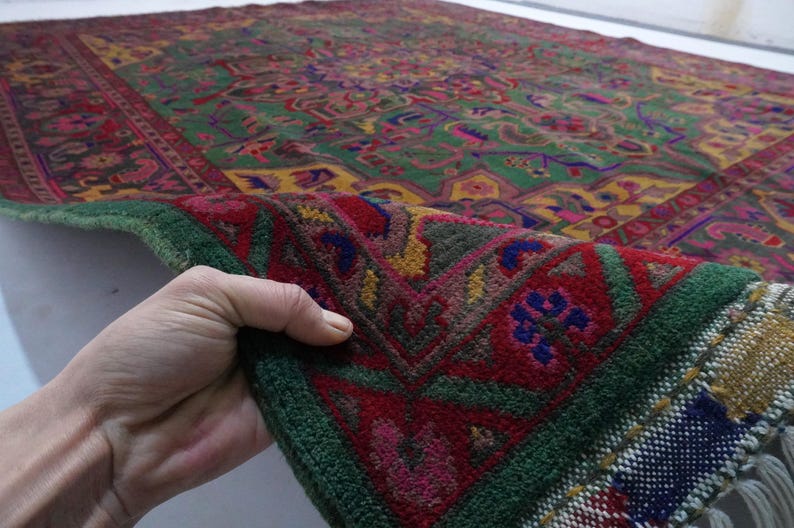 May include: A close-up of a green, red and yellow patterned rug with a fringed border. The rug is folded in half and being held by a hand.