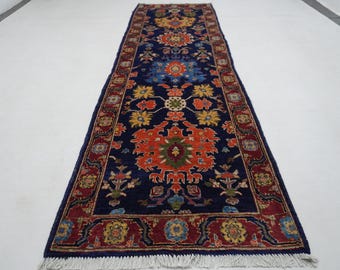 May include: A rectangular, navy blue runner rug with a central floral design in orange, yellow, and green. The rug has a red border with repeating floral patterns and white fringe on the ends. The rug's dimensions are not visible.