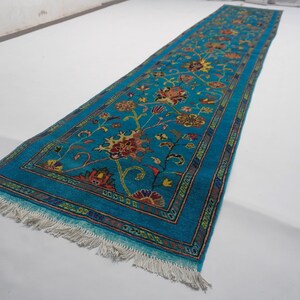 May include: A long, rectangular turquoise rug with a floral pattern. The rug features a central design of flowers in shades of red, yellow, and orange, with green leaves and stems. The border is a darker blue with a repeating pattern. The rug has a fringe on both ends.