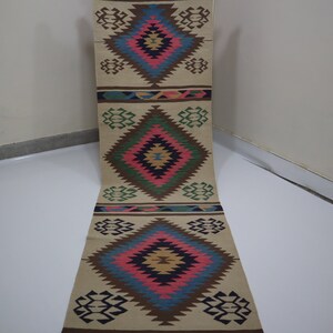 May include: A beige runner rug with a geometric diamond pattern in shades of blue, pink, brown, and green. The rug has a repeating design with diamond shapes and decorative borders. The rug is made of woven material.