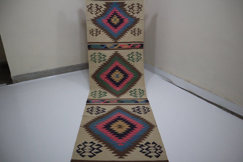 May include: A beige runner rug with a geometric diamond pattern in shades of blue, pink, brown, and green. The rug has a repeating design with diamond shapes and decorative borders. The rug is made of woven material.