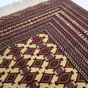 May include: A traditional rug with a cream background and intricate geometric patterns in shades of red, brown, and black. The rug has a fringe border.