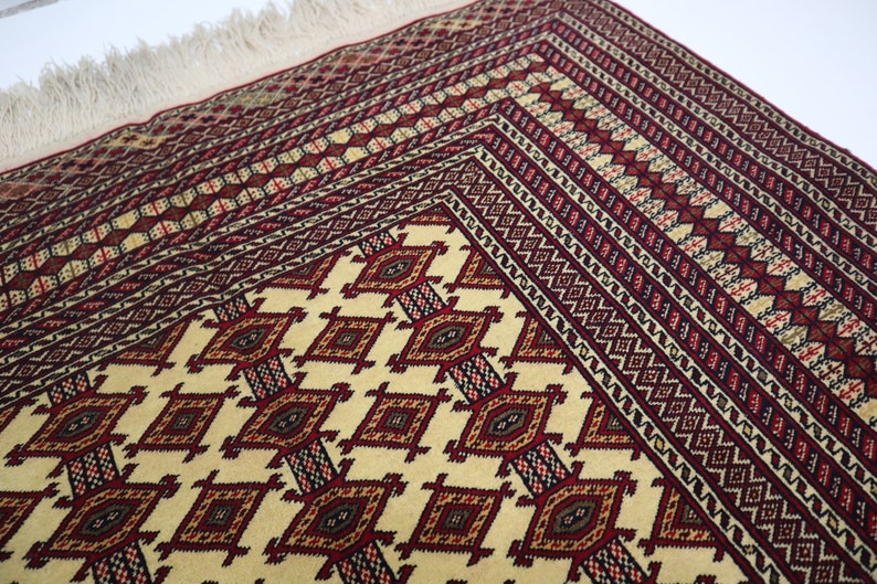 May include: A traditional rug with a cream background and intricate geometric patterns in shades of red, brown, and black. The rug has a fringe border.