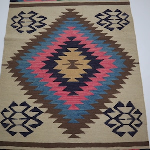 May include: A beige and brown woven rug with a central diamond pattern in pink, blue, and brown. The rug features geometric designs in navy blue at the top and bottom, with a brown border.
