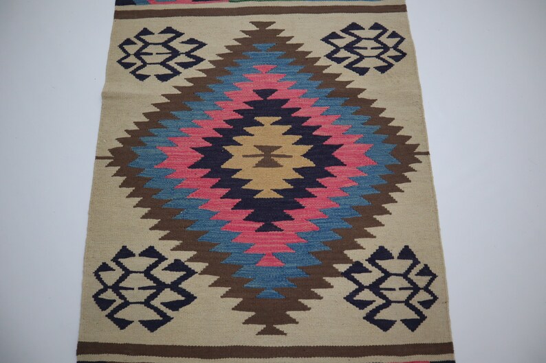 May include: A beige and brown woven rug with a central diamond pattern in pink, blue, and brown. The rug features geometric designs in navy blue at the top and bottom, with a brown border.
