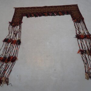 May include: A decorative textile piece with a rectangular top and long, fringed strands. The top features a geometric pattern in shades of brown, with the fringe incorporating red, orange, and cream-colored accents.