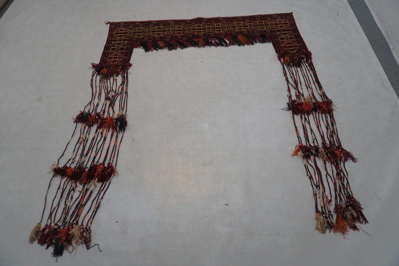 May include: A decorative textile piece with a rectangular top and long, fringed strands. The top features a geometric pattern in shades of brown, with the fringe incorporating red, orange, and cream-colored accents.