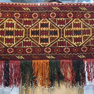 May include: A rectangular, patterned textile with a geometric design in shades of red, gold, and black. The bottom edge features a fringe of various colors, including green, orange, and black, with small white beads.