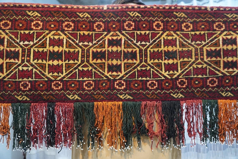 May include: A rectangular, patterned textile with a geometric design in shades of red, gold, and black. The bottom edge features a fringe of various colors, including green, orange, and black, with small white beads.