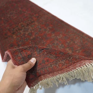 May include: A rectangular runner rug featuring a deep red base color with intricate dark brown and black floral patterns. The rug has a woven texture and is finished with off-white fringe tassels along the short edge, showing the underside.