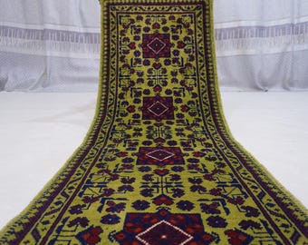 Puede incluir: Una alfombra larga y rectangular con un diseño central de formas geométricas repetidas en rojo, azul y blanco sobre un fondo verde. La alfombra tiene un borde morado oscuro y flecos.