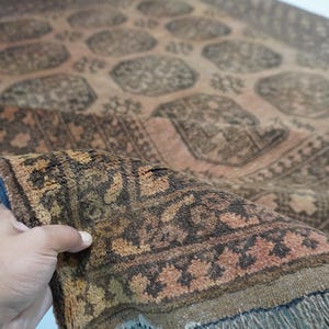 May include: A detailed shot of a vintage rug with a complex geometric pattern in shades of brown, beige, and rust. The rug's edge is folded, revealing a fringe and a dark blue border. The rug appears to be hand-knotted.