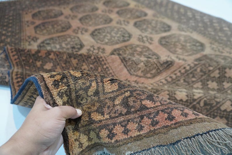 May include: A detailed shot of a vintage rug with a complex geometric pattern in shades of brown, beige, and rust. The rug's edge is folded, revealing a fringe and a dark blue border. The rug appears to be hand-knotted.