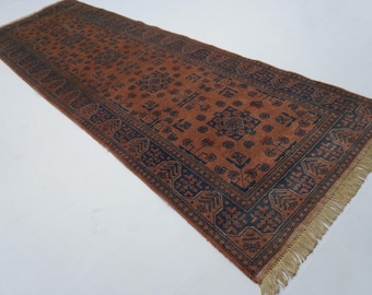 May include: A rectangular, long, and narrow rug with a warm, reddish-brown field and a dark blue border. The rug features a repeating geometric pattern in dark blue and has fringed edges.