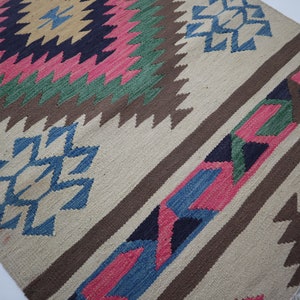 May include: Handwoven rug with a geometric pattern. The rug features a central diamond design in pink, green, and yellow, surrounded by brown and blue accents. The background is a neutral beige color, with brown stripes and blue geometric shapes.