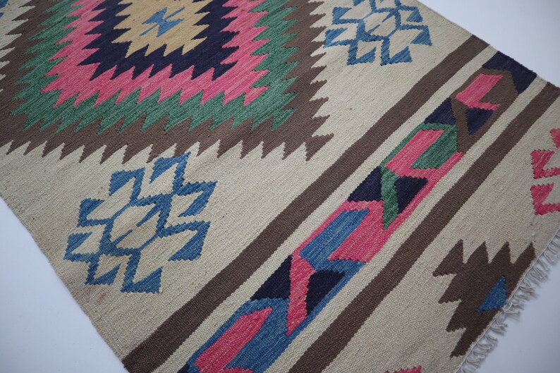 May include: Handwoven rug with a geometric pattern. The rug features a central diamond design in pink, green, and yellow, surrounded by brown and blue accents. The background is a neutral beige color, with brown stripes and blue geometric shapes.