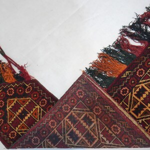 May include: A detailed shot of a traditional rug with a geometric pattern in shades of red, brown, and gold. The rug has decorative tassels in red, orange, and black. The rug is folded, showcasing its intricate design.