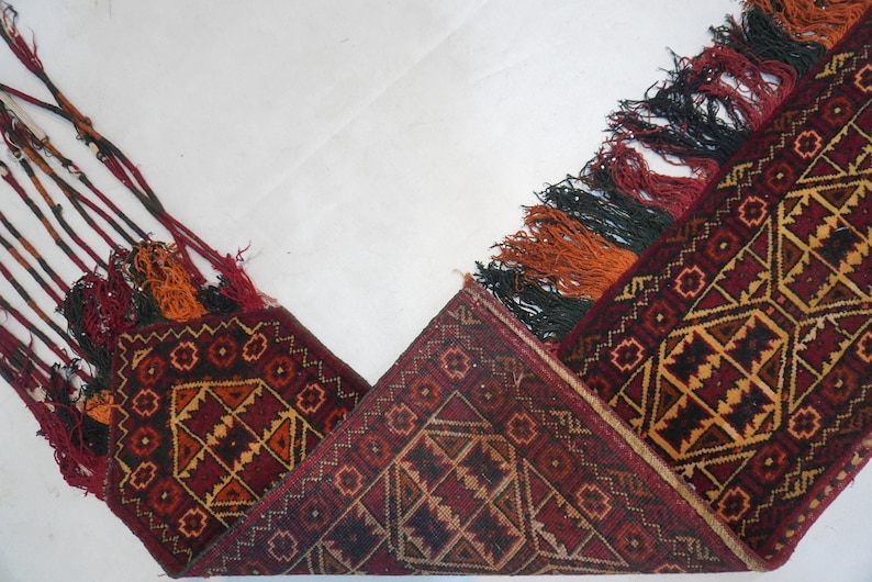 May include: A detailed shot of a traditional rug with a geometric pattern in shades of red, brown, and gold. The rug has decorative tassels in red, orange, and black. The rug is folded, showcasing its intricate design.