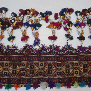 Op de afbeelding: Gedetailleerd textiel met een geometrisch patroon in bruin-, rood- en geeltinten. Het textiel is versierd met kleurrijke kwastjes en decoratieve elementen, waaronder zilverkleurige kegels en kralenaccenten. Het ontwerp heeft een verscheidenheid aan kleuren, waaronder oranje, groen en paars.