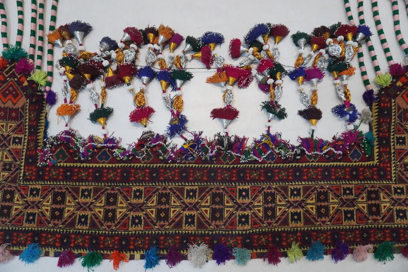 Op de afbeelding: Gedetailleerd textiel met een geometrisch patroon in bruin-, rood- en geeltinten. Het textiel is versierd met kleurrijke kwastjes en decoratieve elementen, waaronder zilverkleurige kegels en kralenaccenten. Het ontwerp heeft een verscheidenheid aan kleuren, waaronder oranje, groen en paars.