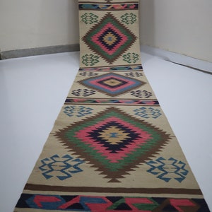 May include: A long, beige runner rug with a repeating geometric pattern in shades of pink, blue, green, and brown. The rug features diamond and zigzag designs, with a fringed edge. The rug is displayed vertically.