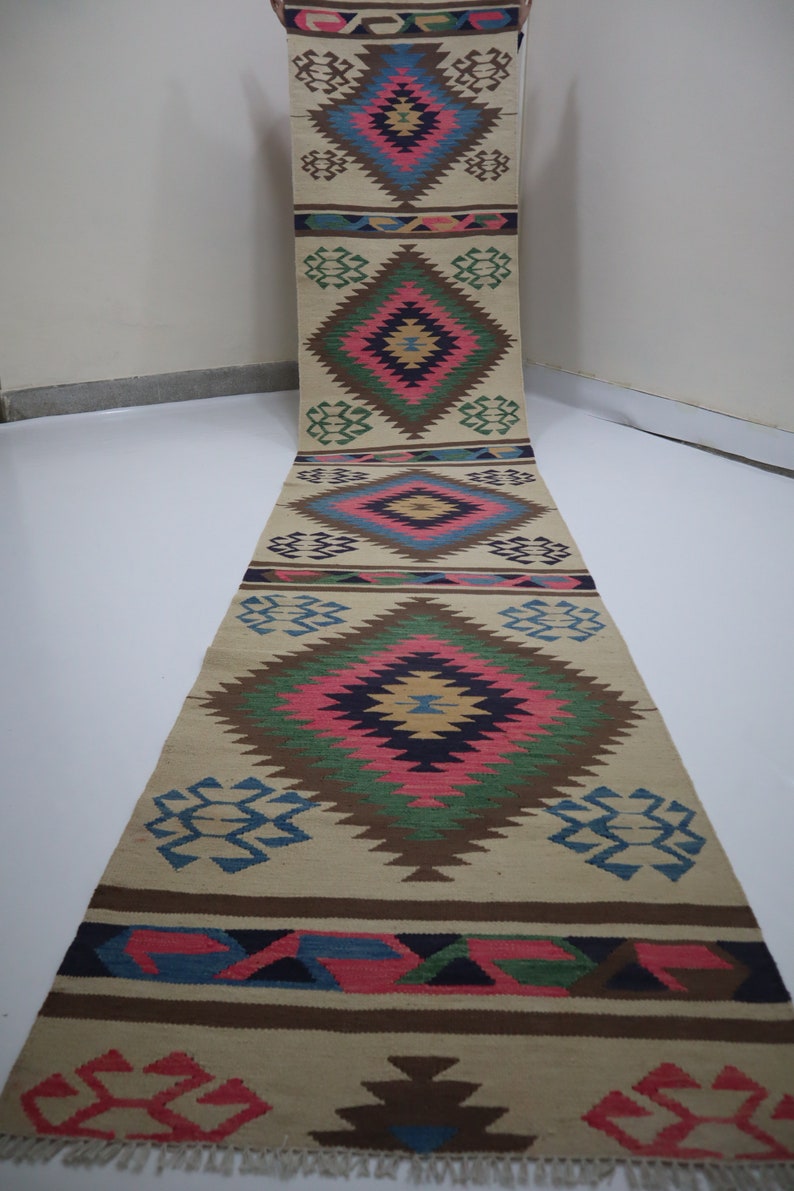 May include: A long, beige runner rug with a repeating geometric pattern in shades of pink, blue, green, and brown. The rug features diamond and zigzag designs, with a fringed edge. The rug is displayed vertically.