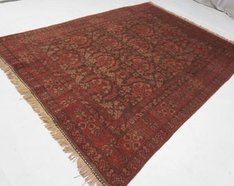 May include: A rectangular area rug featuring a deep red and dark brown color palette with an intricate repeating floral and geometric pattern across the center and a wide decorative border. The rug is finished with light beige fringe on the ends.