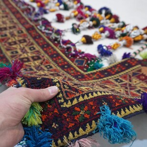 Op de afbeelding: Een hand houdt een deel van een kleurrijk, patroonrijk textiel vast. Het textiel heeft een geometrisch ontwerp in rood, geel en bruin, met een donkerblauwe rand. Kwasten in verschillende kleuren, waaronder blauw, groen en roze, sieren de randen.