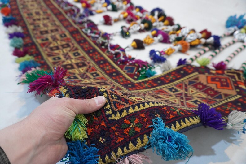 Op de afbeelding: Een hand houdt een deel van een kleurrijk, patroonrijk textiel vast. Het textiel heeft een geometrisch ontwerp in rood, geel en bruin, met een donkerblauwe rand. Kwasten in verschillende kleuren, waaronder blauw, groen en roze, sieren de randen.