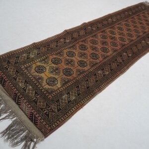 May include: A rectangular runner rug, hand-woven with a central pattern of repeating floral medallions in brown, orange, and tan. The rug has a dark brown border with geometric designs and fringe at each end.