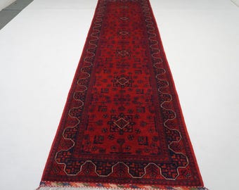 May include: A red runner rug with a traditional Persian design. The rug has a dark blue border with intricate patterns and a red center with a repeating geometric design.