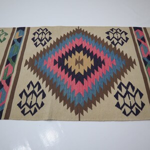 May include: A beige runner rug with a geometric pattern. The design features diamond shapes in pink, blue, brown, and yellow, with accents of green and navy blue. The rug is likely made of woven fabric.