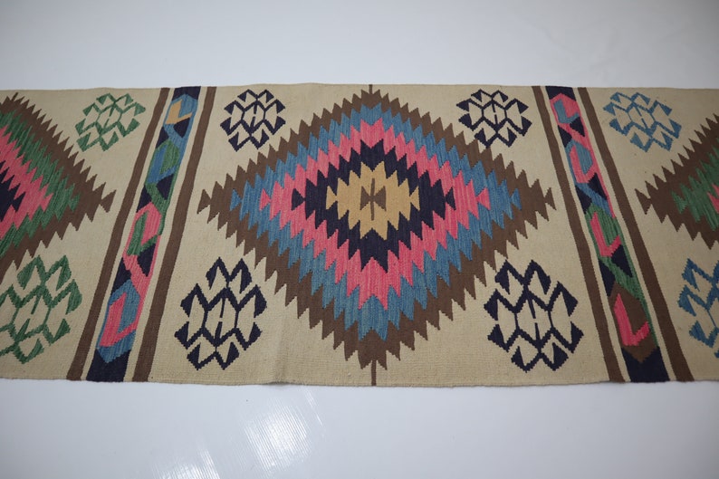 May include: A beige runner rug with a geometric pattern. The design features diamond shapes in pink, blue, brown, and yellow, with accents of green and navy blue. The rug is likely made of woven fabric.