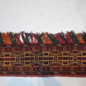 May include: A section of a woven textile with a geometric pattern in shades of red, brown, and gold. The top edge features a row of colorful tassels in red, black, orange, and green. The textile has a detailed, intricate design.