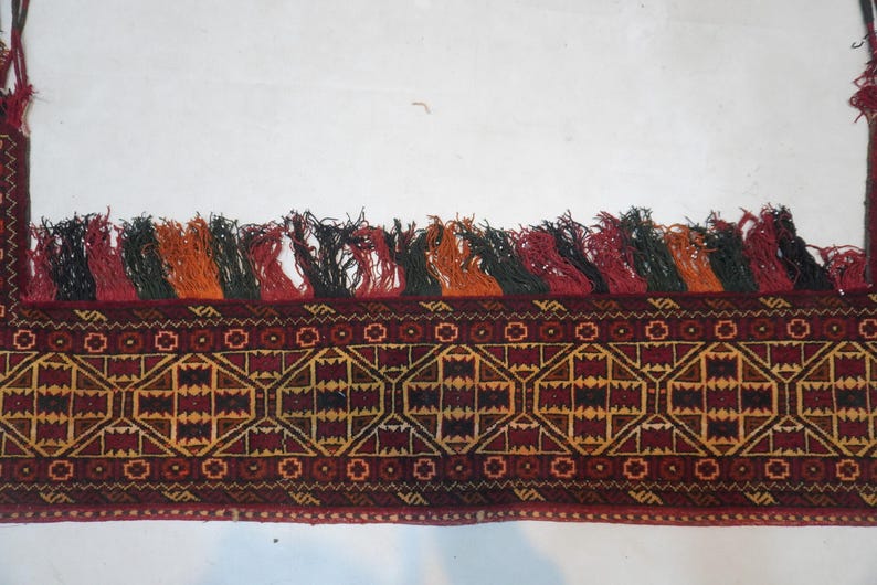 May include: A section of a woven textile with a geometric pattern in shades of red, brown, and gold. The top edge features a row of colorful tassels in red, black, orange, and green. The textile has a detailed, intricate design.