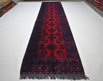 May include: A long, rectangular rug with a deep red central field and intricate dark blue and black floral patterns. The rug has a dark blue border and a gray fringe on both ends. The rug is displayed on a white surface.