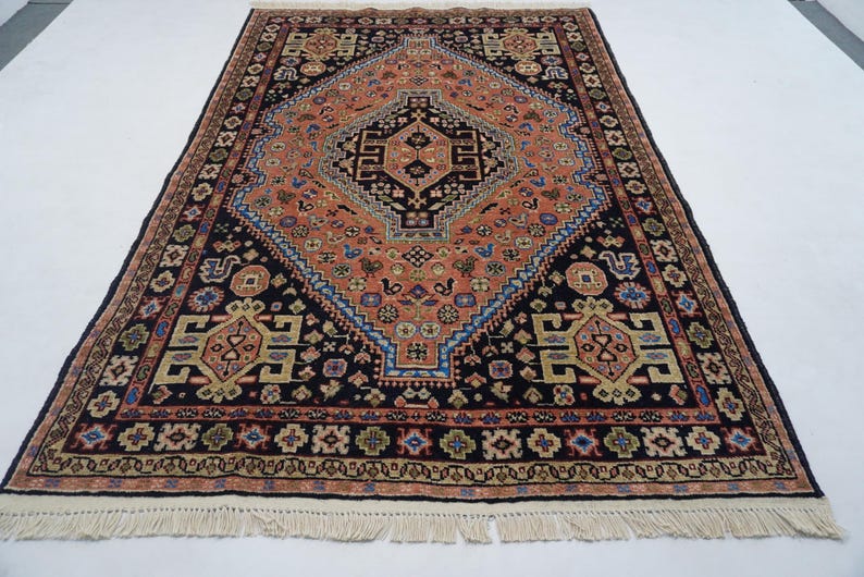 May include: A traditional Persian rug with a black background and intricate patterns in shades of red, orange, blue, and beige. The rug features a central medallion with a diamond shape and a geometric design. The rug has a fringe border.