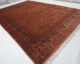 May include: A rectangular area rug featuring a deep terracotta and rust-colored field with intricate dark brown geometric patterns and floral motifs. The rug is framed by a wide, detailed border and finished with short white fringe on the ends.